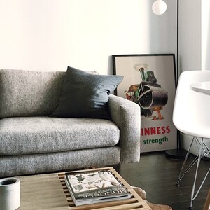 May include: A modern living room scene featuring a gray sofa with a dark gray pillow. A framed Guinness poster is visible, along with a white chair and a small round table. A coffee table with a magazine and a concrete pot completes the decor.