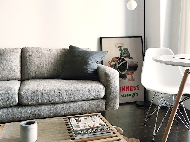 May include: A modern living room scene featuring a gray sofa with a dark gray pillow. A framed Guinness poster is visible, along with a white chair and a small round table. A coffee table with a magazine and a concrete pot completes the decor.