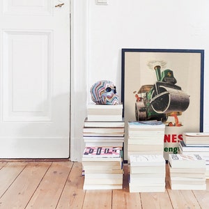 May include: A colorful skull sculpture sits atop a stack of books, next to a framed vintage poster of a steamroller. Several stacks of books are arranged on a wooden floor, creating a decorative display. The poster has the word "NES" visible.