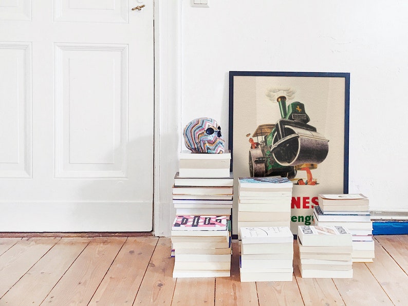 May include: A colorful skull sculpture sits atop a stack of books, next to a framed vintage poster of a steamroller. Several stacks of books are arranged on a wooden floor, creating a decorative display. The poster has the word "NES" visible.