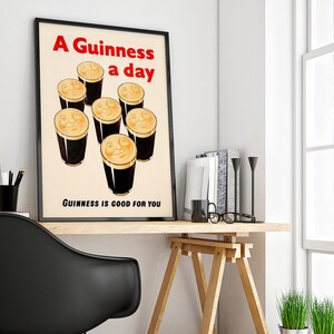 May include: A framed vintage poster featuring the text "A Guinness a day" in red, above cartoon pints of Guinness with smiling faces. The text "Guinness is good for you" is below. The poster is on a wooden desk, with books and a black chair.