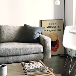 K&ouml;nnte beinhalten: Ein modernes Wohnzimmer mit einem grauen Sofa und einem dunkelgrauen Kissen. Ein gerahmtes Guinness-Poster mit dem Text "Have a Guinness when you're tired" lehnt an der Wand. Ein wei&szlig;er Stuhl und ein kleiner Tisch sind ebenfalls zu sehen.