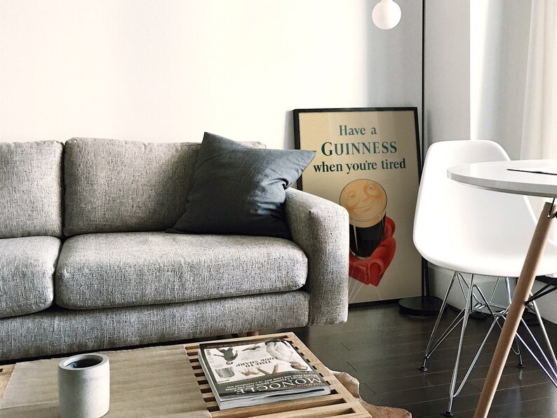 K&ouml;nnte beinhalten: Ein modernes Wohnzimmer mit einem grauen Sofa und einem dunkelgrauen Kissen. Ein gerahmtes Guinness-Poster mit dem Text "Have a Guinness when you're tired" lehnt an der Wand. Ein wei&szlig;er Stuhl und ein kleiner Tisch sind ebenfalls zu sehen.