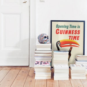 May include: A framed poster with the text "Opening Time is Guinness Time" featuring a toucan with a Guinness bottle. A colourful skull and several stacks of books are in front of a white door and wooden floor.