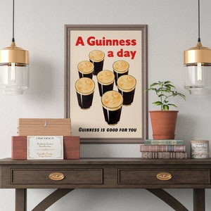 May include: A framed vintage Guinness poster with seven smiling pints of stout. The red text reads "A Guinness a day", with "Guinness is good for you" below. The poster is displayed on a wooden table with decorative items, including a plant and books.
