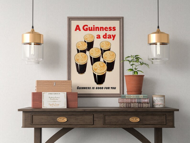 May include: A framed vintage Guinness poster with seven smiling pints of stout. The red text reads "A Guinness a day", with "Guinness is good for you" below. The poster is displayed on a wooden table with decorative items, including a plant and books.