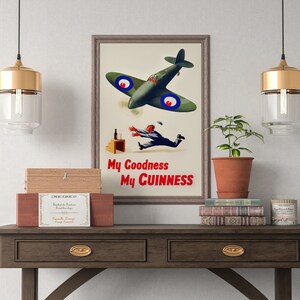 May include: Framed vintage advertisement poster with a green airplane, a falling man, and the text "My Goodness My Guinness." The poster is displayed on a wooden table with decorative items, including a plant, books, and a wooden box.