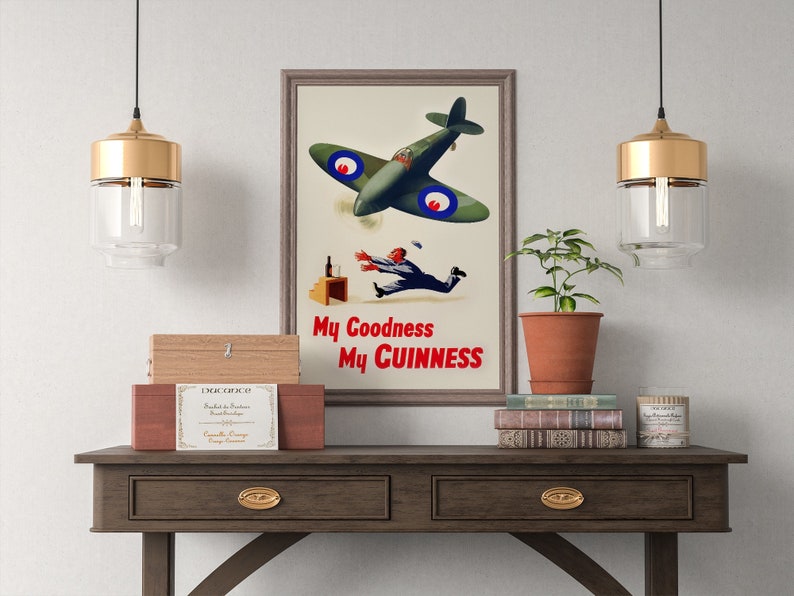 May include: Framed vintage advertisement poster with a green airplane, a falling man, and the text "My Goodness My Guinness." The poster is displayed on a wooden table with decorative items, including a plant, books, and a wooden box.