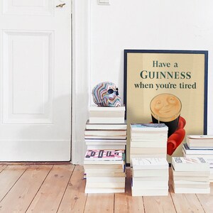 K&ouml;nnte beinhalten: Ein gerahmtes Guinness-Poster mit dem Text "Have a Guinness when you're tired" lehnt an einer wei&szlig;en Wand. Ein bunter Totenkopf sitzt auf einem Stapel B&uuml;cher. Mehrere Stapel B&uuml;cher stehen auf dem Holzboden.
