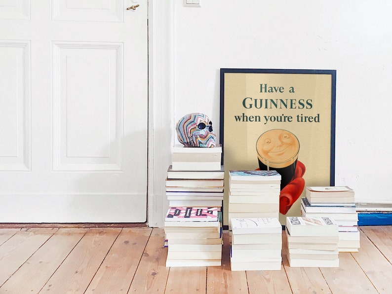 K&ouml;nnte beinhalten: Ein gerahmtes Guinness-Poster mit dem Text "Have a Guinness when you're tired" lehnt an einer wei&szlig;en Wand. Ein bunter Totenkopf sitzt auf einem Stapel B&uuml;cher. Mehrere Stapel B&uuml;cher stehen auf dem Holzboden.