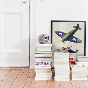 May include: A colorful skull sits atop a stack of books next to a framed vintage aviation poster. The poster features a green airplane and a falling figure. Several stacks of books are arranged on a wooden floor.