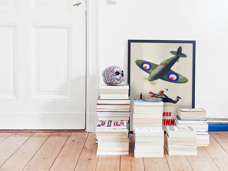 May include: A colorful skull sits atop a stack of books next to a framed vintage aviation poster. The poster features a green airplane and a falling figure. Several stacks of books are arranged on a wooden floor.