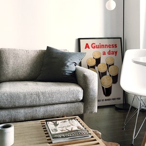 May include: A modern living room with a grey sofa and a dark grey cushion. A framed poster with the text "A Guinness a day is good for you" is visible. A white table and chair are also present, along with a coffee table with a magazine and a small concrete pot.