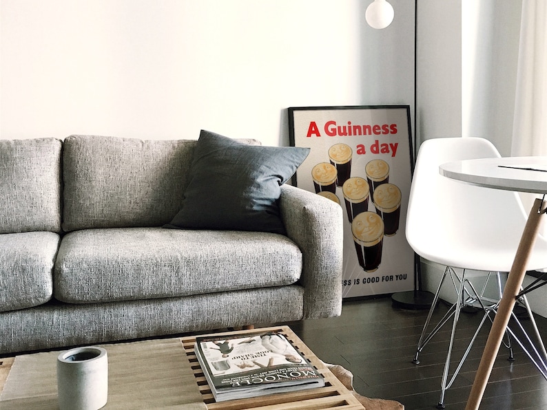 May include: A modern living room with a grey sofa and a dark grey cushion. A framed poster with the text "A Guinness a day is good for you" is visible. A white table and chair are also present, along with a coffee table with a magazine and a small concrete pot.