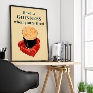 K&ouml;nnte beinhalten: Gerahmtes Poster mit dem Text "Have a GUINNESS when you're tired". Das Bild zeigt ein Pint Guinness Stout mit einem l&auml;chelnden Gesicht, das auf einem roten Sessel ruht. Das Poster steht auf einem Holztisch mit B&uuml;chern und einem schwarzen Stuhl.