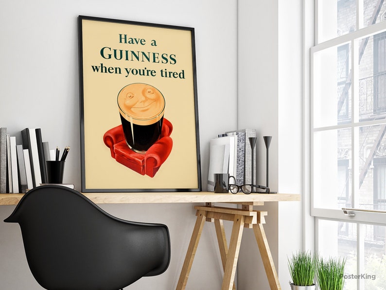 K&ouml;nnte beinhalten: Gerahmtes Poster mit dem Text "Have a GUINNESS when you're tired". Das Bild zeigt ein Pint Guinness Stout mit einem l&auml;chelnden Gesicht, das auf einem roten Sessel ruht. Das Poster steht auf einem Holztisch mit B&uuml;chern und einem schwarzen Stuhl.