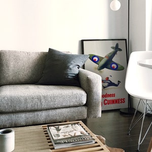 May include: A living room scene featuring a gray sofa with a dark gray pillow. A framed vintage poster with a green airplane and the text "Goodness Guinness" leans against the wall. A white chair and a round table are also visible.