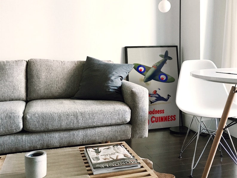 May include: A living room scene featuring a gray sofa with a dark gray pillow. A framed vintage poster with a green airplane and the text "Goodness Guinness" leans against the wall. A white chair and a round table are also visible.