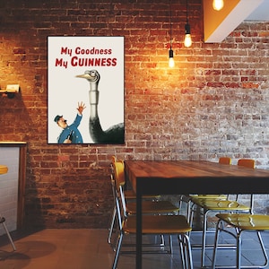 May include: A framed vintage advertisement poster with the text "My Goodness My Guinness" in red. The poster features a cartoon illustration of a man in a blue uniform reaching up towards a large bird-like creature. The background is a brick wall.