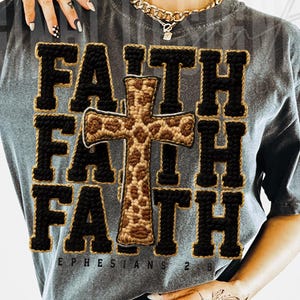 May include: Gray t-shirt with the word "FAITH" in a bold, black, and gold design. A cross with a leopard print pattern is centered on the shirt. The text "EPHESIANS 2:8" is printed at the bottom.
