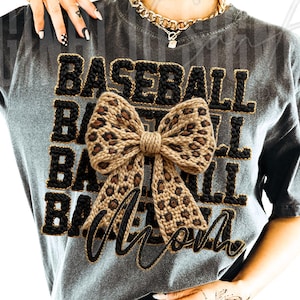 May include: Gray t-shirt with the words "BASEBALL MOM" in black and gold lettering. A large bow with a leopard print design is centered on the shirt. The shirt has short sleeves and a crew neck.