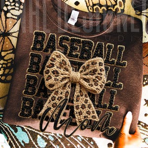 May include: A brown sweatshirt with the word "BASEBALL" in black, block letters. A leopard print bow is centered on the shirt. The word "MOM" is embroidered in black script below the bow. The shirt is displayed on a patterned surface.