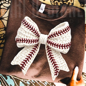 May include: A brown sweatshirt with a large, white, baseball-themed bow. The bow is made of a knitted material with red stitching, resembling the seams of a baseball. The bow is centered on the sweatshirt.