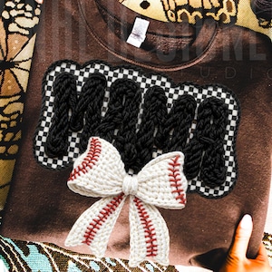 May include: A brown sweatshirt with the word "MAMA" in black, textured letters outlined with a black and white checkered pattern. A white crocheted bow with red stitching, resembling a baseball, is centered below the text.