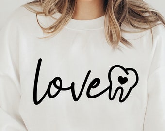 May include: A white sweatshirt with the text "love" in black cursive lettering and a black tooth with a small red heart inside. The model is wearing a white beanie and has long brown hair.