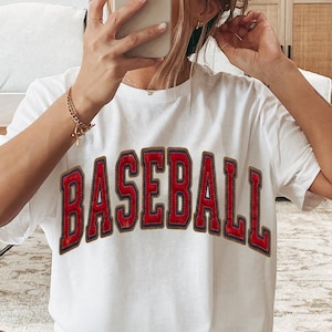 Peut inclure: T-shirt blanc avec le mot "BASEBALL" en grandes lettres rouges cousues. Les lettres ont un contour doré. Le t-shirt est de style décontracté, à manches courtes.