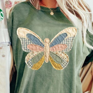 May include: A green t-shirt featuring a patchwork butterfly design. The butterfly has wings in various colors and patterns, including blue, pink, and yellow. The text "ORIGINAL DESIGNER CANARTSTUDIO" is printed at the bottom right.