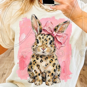Op de afbeelding: Crèmekleurig T-shirt met een luipaardprint konijn met een roze strik. Het konijn staat tegen een roze geschilderde achtergrond. Het shirt heeft een ronde hals en korte mouwen. Het woord "STUDIO" is gedeeltelijk zichtbaar.