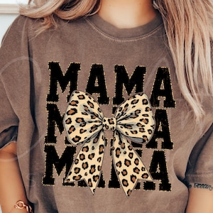Puede incluir: Una camiseta marrón con el texto "MAMA" impreso en negro y un gran lazo de estampado de leopardo en el centro.