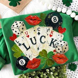 Puede incluir: Sudadera verde con un diseño que presenta naipes que deletrean "LUCK", dados, labios rojos, corazones y una bola ocho negra. El diseño también incluye tréboles. Tema del Día de San Patricio.