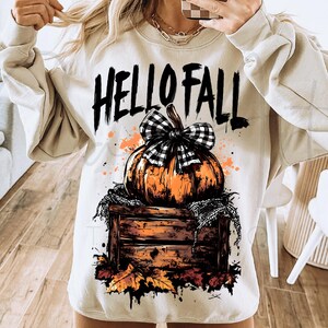 May include: Beige sweatshirt with a fall-themed graphic. The design features an orange pumpkin with a black and white checkered bow, set on a wooden crate with autumn leaves. The words "HELLO FALL" are printed above the pumpkin.