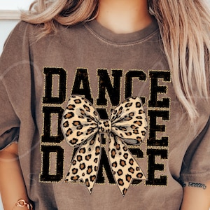 May include: A brown t-shirt featuring the word "DANCE" in a black, blocky font. A large bow with a leopard print design is centered on the shirt. The shirt has short sleeves.