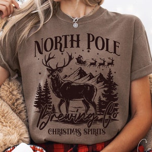 May include: A brown t-shirt with a graphic design of a deer, mountains, and the text "North Pole Brewing Co. Christmas Spirits".