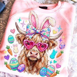 Puede incluir: Sudadera rosa claro con una vaca de las Tierras Altas que lleva gafas de sol en forma de corazón y orejas de conejo. La vaca está rodeada de huevos de Pascua, zanahorias y otras decoraciones festivas. Un lazo colorido adorna la cabeza de la vaca.