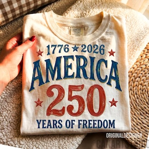 Può includere: T-shirt color crema con la scritta "1776-2026 AMERICA 250 YEARS OF FREEDOM" in blu e rosso. La maglietta ha un girocollo e una piccola etichetta con la scritta "Comfort Colors". La maglietta è piegata.