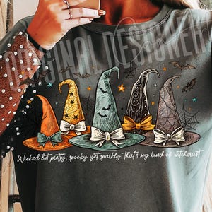 May include: Dark gray t-shirt featuring five witch hats in orange, yellow, teal, black, and gray, each with a bow. Bats, spiderwebs, and stars decorate the design. Text reads: "Wicked But pretty, spooky yet sparkly. That's my kind of witchcraft."