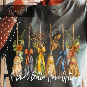 以下が含まれることがあります： 6本の装飾的なほうきのデザインが施された濃い灰色のTシャツ。それぞれカラフルなリボンと花の装飾が施されています。「A Girl Gotta Have Options」の文字がほうきの下に白い筆記体で印刷されています。