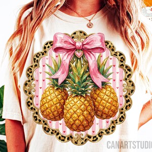 May include: Cream-colored t-shirt featuring a pineapple design. The design includes three golden pineapples, a pink bow, and a heart-shaped accent. The design is set within a scalloped frame with a leopard print border.
