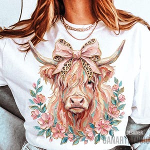 May include: White t-shirt featuring a colorful illustration of a Highland cow with a pink bow and floral wreath. The cow has long, shaggy fur in shades of pink, peach, and cream. The bow has a leopard print pattern.