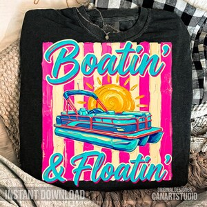 May include: Dark gray long-sleeve shirt with a colorful graphic. The graphic features a blue and pink pontoon boat with the words "Boatin' & Floatin'" in turquoise script. A yellow sun is in the background.