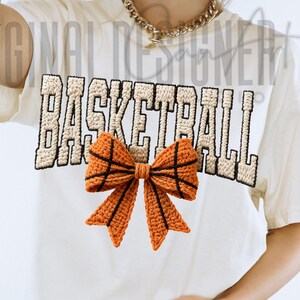 Op de afbeelding: Cr&egrave;mekleurig T-shirt met het woord "BASKETBALL" in een beige, getextureerde lettertype. Onder de tekst bevindt zich een oranje en zwarte gehaakte strik, die op een basketbal lijkt. Het shirt heeft een casual, sportief ontwerp.
