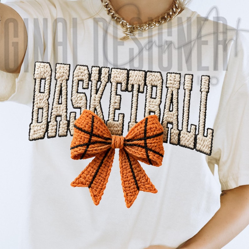 Op de afbeelding: Cr&egrave;mekleurig T-shirt met het woord "BASKETBALL" in een beige, getextureerde lettertype. Onder de tekst bevindt zich een oranje en zwarte gehaakte strik, die op een basketbal lijkt. Het shirt heeft een casual, sportief ontwerp.