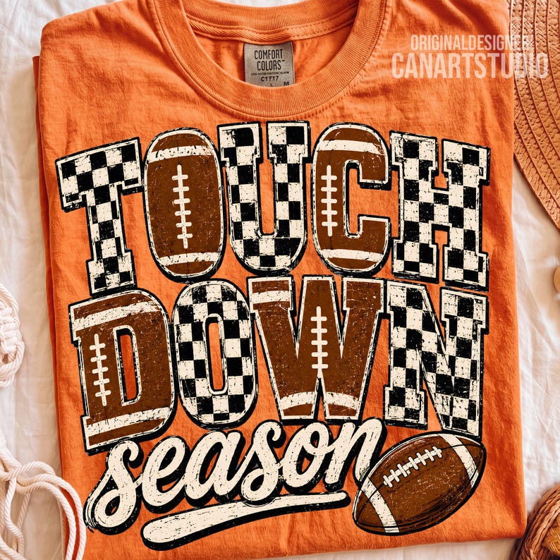 May include: An orange t-shirt featuring the text TOUCH DOWN SEASON in a distressed font. The letters are filled with a black and white checkered pattern and brown football graphics. A brown football illustration is positioned at the bottom right.