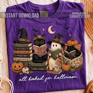 May include: A purple t-shirt featuring a graphic of a black cat and a ghost wearing witch hats, reading books. The design includes stacks of books, pumpkins, a skull, and a crescent moon. The text reads "all booked for halloween".