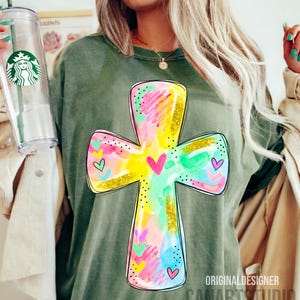 Op de afbeelding: Olijfgroen T-shirt met een kleurrijk kruisontwerp. Het kruis is versierd met aquarel-achtige spetters in roze, geel, blauw en groen, geaccentueerd met gouden glitter en hartdetails. De tekst "ORIGINAL DESIGNER CANARTSTUDIO" staat onderaan.