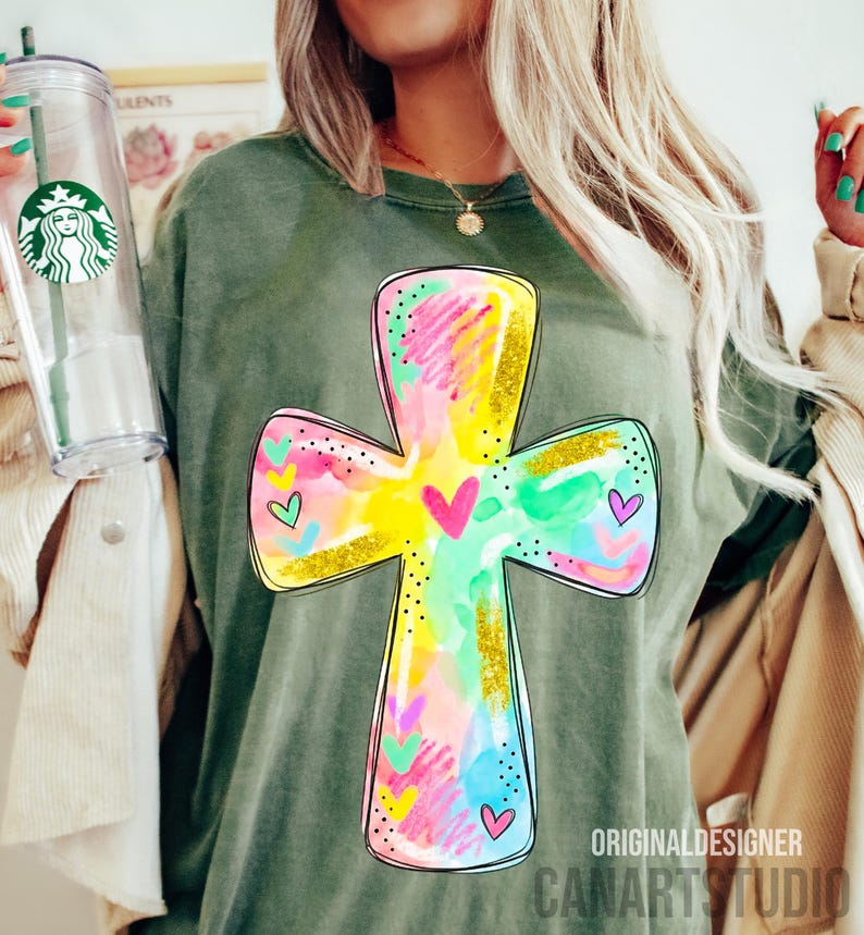 Op de afbeelding: Olijfgroen T-shirt met een kleurrijk kruisontwerp. Het kruis is versierd met aquarel-achtige spetters in roze, geel, blauw en groen, geaccentueerd met gouden glitter en hartdetails. De tekst "ORIGINAL DESIGNER CANARTSTUDIO" staat onderaan.